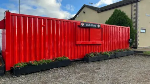 A large red metal shipping container. It has a close hatch in the middle and sign saying 