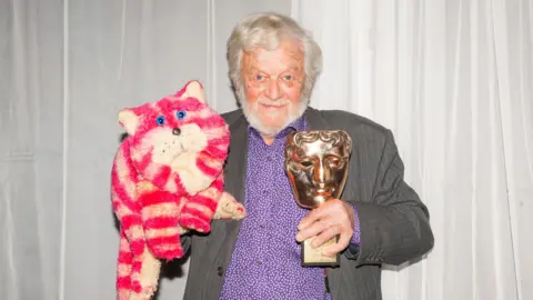 Dominic Lipinski/PA Wire A man with grey hair, purple shirt and grey suit holding a Bafta trophy and a cuddly Bagpuss doll.