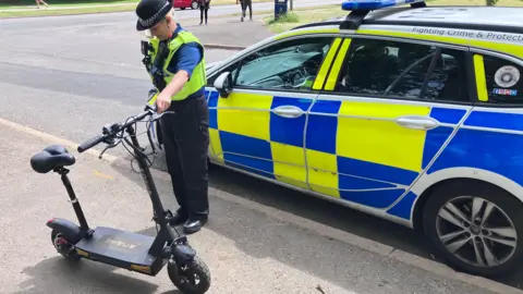 Sam Read/BBC A female police officer stands on the pavement next to a police car, holding a black e-scooter. She is wearing a black uniform and a black police hat.