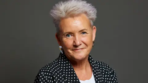 Parliamentary portrait of Lorraine Beavers. She smiles in front of a grey backdrop. She has spiky white hair and a polka dot top. 