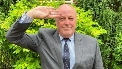 Local Democracy Reporting Service A man wearing a grey suit, blue and white shirt and dark blue patterned tie, smiling and saluting the camera
