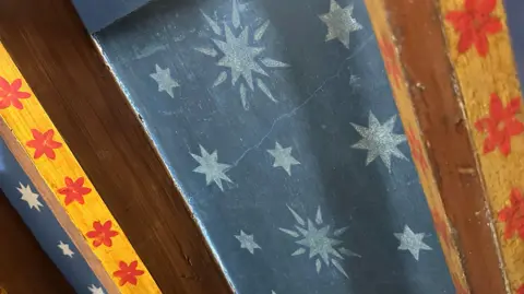 A close up of Pugin's original work which is a dark blue base layer of paint with faded blue stars. Along the beams are red flowers. 