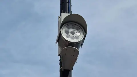 BBC/Naj Modak A close-up of a bus lane enforcement camera mounted on a tall black pole, showing a circular arrangement of multiple light modules beneath a curved protective hood. The fixture includes visible wiring running down the pole. The background is a soft, overcast sky with thin clouds. 