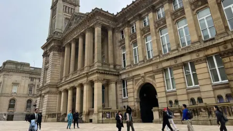 A photo of Birmingham Museum and Art Gallery. It is a building made out of sand coloured stone and there are 10 people walking past in the photo walking in both directions. 