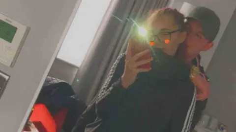 Jasmin Mae Grindley A man and a woman stand close together in front of a full-length mirror inside a bedroom. One of them is holding a phone with the flash on, creating a bright reflection in the mirror. There is a snapchat filter on the image causing emojis to appear next to the girl's face. Both individuals are dressed casually; one is wearing a black jacket with white stripes on the sleeves and blue jeans, while the other is in dark clothing and a cap. The background shows a grey curtain, and a bedside table with items on it. A red bag is visible on a chair near the wall.