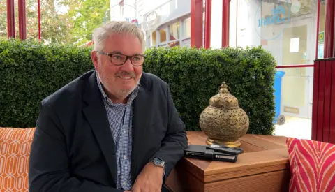 David Voisin: A man with white hair, black rectangle glasses, grey/white stubble, smiling with teeth, wearing a navy suit with a blue chequered shirt with white buttons, a silver watch on his left wrist. He is sat with his left arm resting on a wooden ledge, with his black phone and black notebook beside, a golden ornament behind it. To his right is an orange patterned pillow, behind him is a fake long green bush, windows looking out onto the high street.