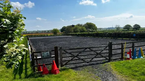 Amanda White / BBC The riding paddock at Kesteven Rideability. It has a black surface and is surrounded by a fence and a hedge.