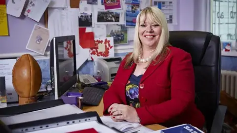 Joy Ballard a blonde haired woman, sat at her desk and smiling at the camera, wearing a red jacket and dress.