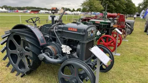 Stuart Howells/BBC Old tractors are lined up within an area of the Suffolk Show, coloured in a mixture of grey, racing green and red. They each have an information board in front of them detailing their history.