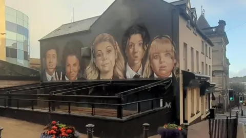 Mural da BBC Derry Girls mostrando os personagens em uma parede de empena com um toldo bloqueando a visão da metade inferior