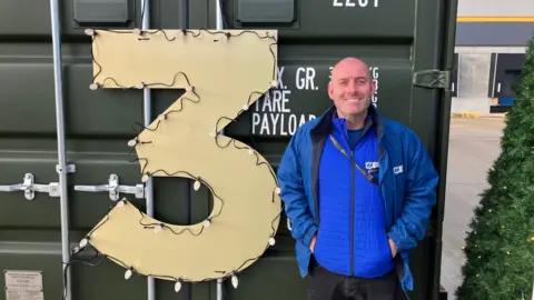 A man in a blue gilet and blue coat stands next to a shipping container. On the front of the container is a a giant number three, covered in lights.