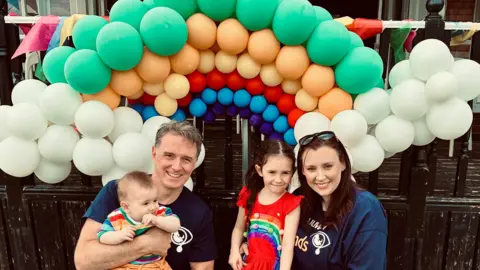 Emily Brazier A man with brown hair is holding a baby who is is weaing a rainbow top and orange shorts and chewing his hand.  A woman with brown hair and sunglasses  on her head is next to a young girl with long brown hair who is wearing a red dress with a rainbow on it. They are stood in front of balloons in a rainbow shape.