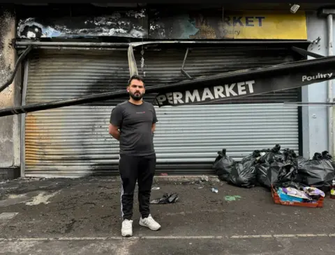 Pacemaker Abdelkader standing in front of his store where the signage has been destroyed by fire and bags of rubbish are visible in front