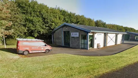 Google A Google street view image of a small office building with a red van parked next to it. A white sign which says "Gilks" can be seen on the building. 