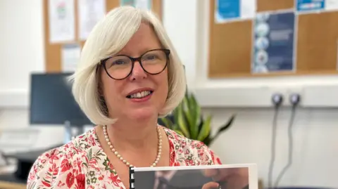 Dr Nicola Brink holding the Public Health's Combined Substance Use Strategy 2025 Annual Report in her office. She has medium length white hair and glasses, with a floral dress on.