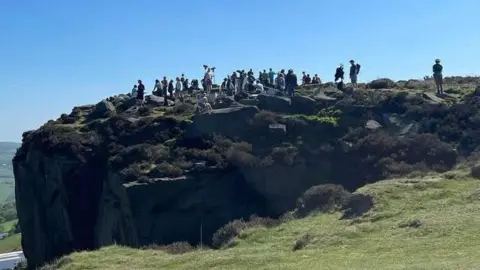 A large group of people on top of the rocks of Ilkley Moor, seen from a distance.