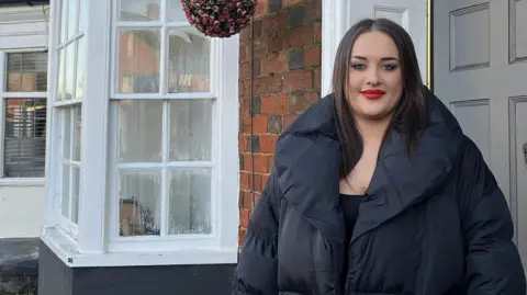 Ellie stands outside her front door which is grey amid traditional brickwork. The row of houses continues behind her, with white bay windows and a sign for the New Inn pub in the distance. Ellie has bright red lipstick and wears a big navy puffer coat. She has straight dark brown hair.