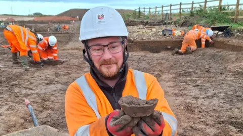 Archaeological Research Services A man in high vis clothing holding a piece of muddy pottery