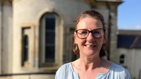 Deirdre Goodlad berdiri di depan Gereja Ramoa Irlandia di Ballycastle. Matahari merenungkan bangunan di belakangnya. Deirdre mengenakan atasan biru dan kacamata. Dia tersenyum dan melihat langsung ke kamera.