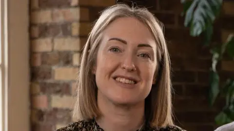 Esther Ghey, who has straight, shoulder length blonde hair, smiles for the camera. She is photographed in front of a brick wall and a large green plant.