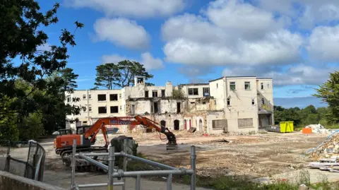 The St Martin's Hotel, a big yellow building that has been partially demolished. In front of it is an orange digger with the words Bob Froome on it. 