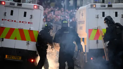 Getty Images  PSNI officers in riot gear responding to unrest. Sparks are close to the officers. There's a large crowd in front of them. The officers have shields and are behind police jeeps. 