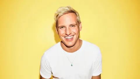 Jamie Laing smiles in front of a yellow background - he has short blond hair and wears a white T-shirt 