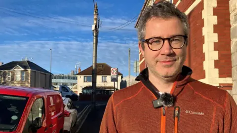 A man wearing a brown and red fleece top, and round glasses. He is smiling at the camera. In the background can be seen a residential street with a parked Royal Mail van and other parked vehicles.