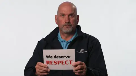 Essex County Council A recycling centre worker sitting in front of a white background. He has a shaved head and a goatee. He is wearing a black jumper over a blue polo top. He is holding a sign that reads "we deserve respect".