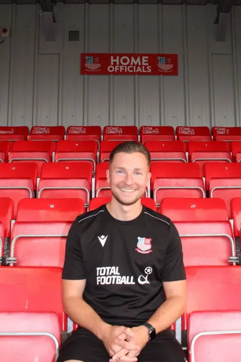 Bowers and Pitsea FC Manager Joe Flower is pictured sitting in the stands at the club's home stadium, the Len Salmon Stadium. The banner directly behind him labels the red seating area for "Home Officials"