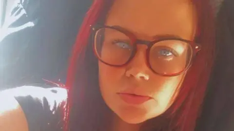 PA Media Holly Sanchez, who has long red hair and blue eyes, wears large glasses as she stares into the camera