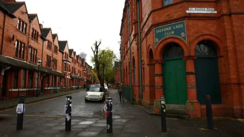 Reuters A street view of the outside of Salford Lads Club, a red-tiled building with a green sign with embossed gold lettering. A man can be seen walking passed on an overcast day.