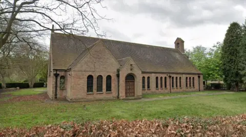 Google A single storey church building with a grassy lawn and trees.