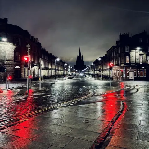 RIP Dornan A dark Edinburgh street view, with red traffic lights reflected on the cobbled street and pavement, and a cathedral silhouette in the distance.