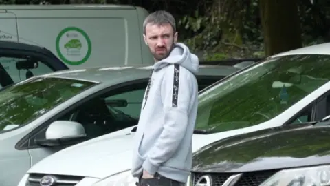 A man with brown hair and beard, wearing a grey hoodie. He is stood in front of cars and looking off to the side with a frown.