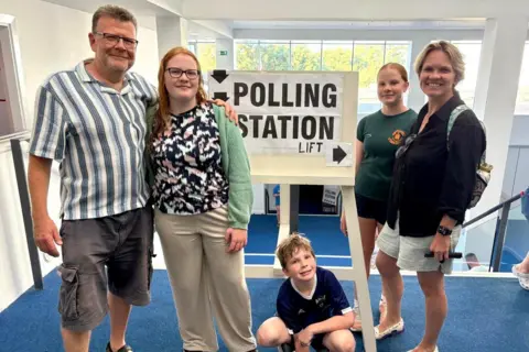 Sasha Kazantseva-Miller A middle-aged man with short brown hair and glasses is looking at the camera and smiling. His arms is round a young woman with long ginger hair and glasses who is looking at the camera and smiling. There is a sign saying 'polling station' beside them. A young boy crouching down beneath the sign and looking at the camera smiling. A young girl with tied-back ginger hair is looking at the camera and smiling, next to a woman with short blonde hair looking at the camera and smiling.