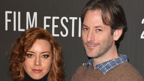 Aubrey Plaza and Jeff Baena smiling in front of a black backdrop, on which the words "film festival" are written