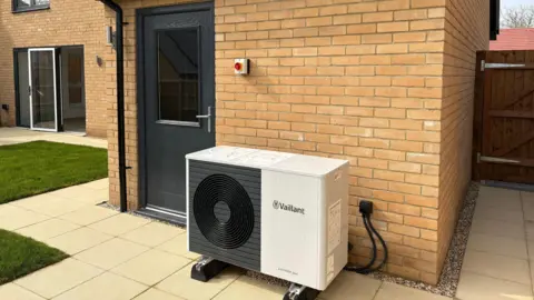 PA Media The back garden of a new house with an external air source heat pump plugged in on the patio. It is white and grey with a Valliant logo. 
