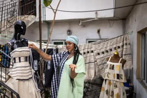 Olympia de Maismtt / AFP stands a woman in a courtyard surrounded by the dresses she designed. They are pointed and mostly neutral tones.