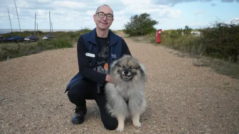 Shaun Whitmore/BBC Man with bald head and glasses and dark jacket with a fluffy grey and brown medium-sized dog with cream-coloured legs.