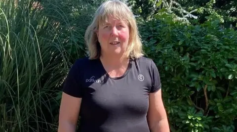 Jenny Stafford is smiling at the camera and her guide dog Nan. She is wearing a black running top with pink shorts. In the background is greenery in a garden.