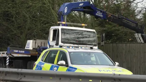 Martin Giles/BBC A yellow and blue police car can be seen on a carriageway. Behind it is a white and blue recovery vehicle. 