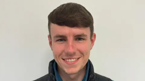 Jack Neill White wall background, shoulders up, Jack is smiling looking directly at the camera. He is wearing a blue fleece under a black waterproof looking coat. He has short brown hair. 