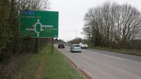 Google A Google street view image of a main road with a large green sign on the left denoting a roundabout. Cars can be seen driving both ways.