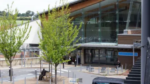 An artists impression of Queen's Centre at Castle Hill Hospital. It is a modern building with open-styled windows and a road going round a bench and trees at the front with a few people sat down.