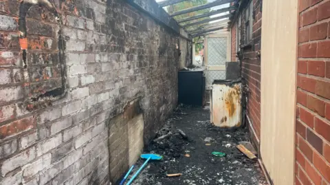 A wall can be seen on one side of the house with what was thought to be a lean to missing its roof with short burnet beams visible. There are charred remains and glass on the floor.