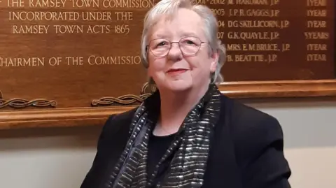 Moira Chrystal Moira Chrystal, who has short grey hair and glasses and is standing in Ramsey Commissioners offices.