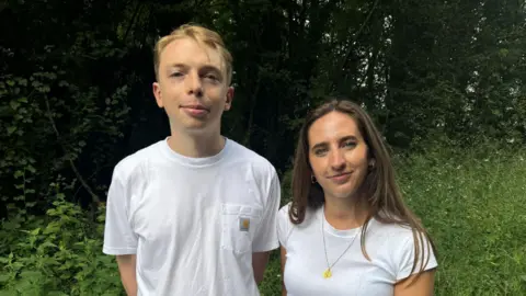 Hannah Roe/BBC Two young, white people. One is male, on the left, the other is female. The man on the left has blonde hair and is wearing a white t-shirt. The woman on the right has long brown hair. Both are smiling and looking at the camera. They are stood in front of green hedgerow.