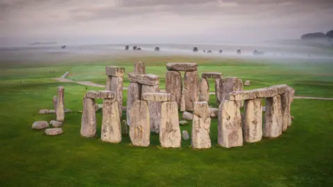 The image shows Stonehenge. There is a misty sky behind with the full circles of the ancient monument in the foreground. Four sets of two large upright stones, each with another large stone resting across the top, which make up part of the outer circle. The even larger stones of the inner area are visible above those of the outer circle. Green grass surrounds the monument on all sides. 