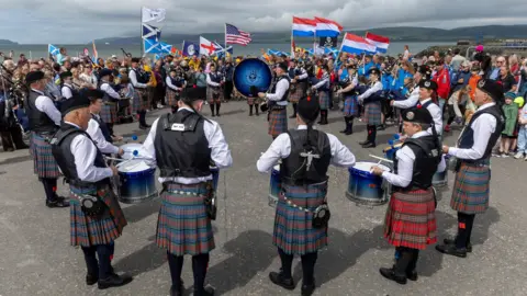 Colin Tennant CT Productions Members of a pipe band standing outside at Straraer harbour. The drummers and pipers are standing in a circle as they preform a tune. A crowd, with some people holding flags of different countries, looks on. The sea and distant shore are in the background.